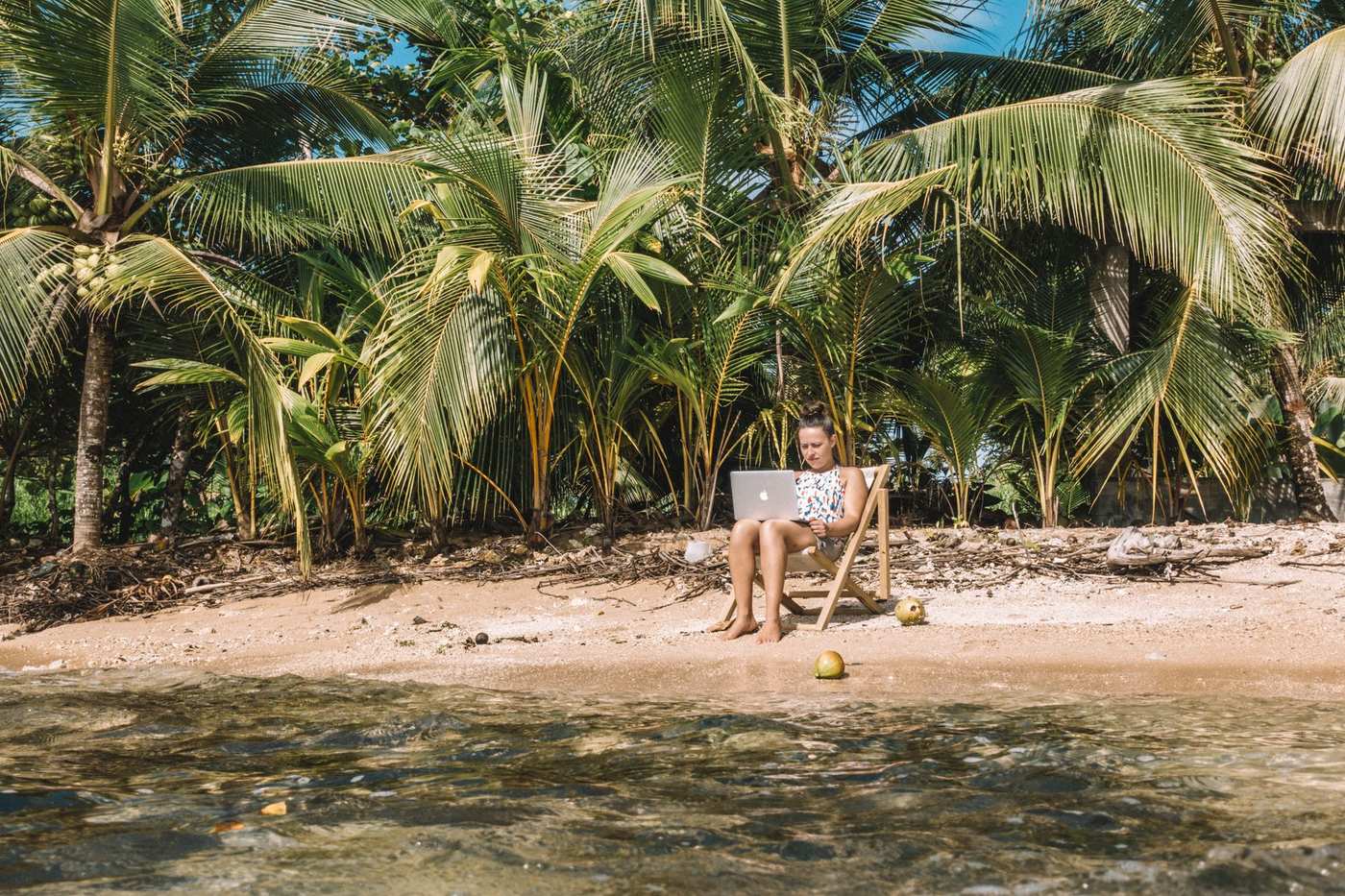 Woman working on a laptop in a beach chair beside palm trees and clear water