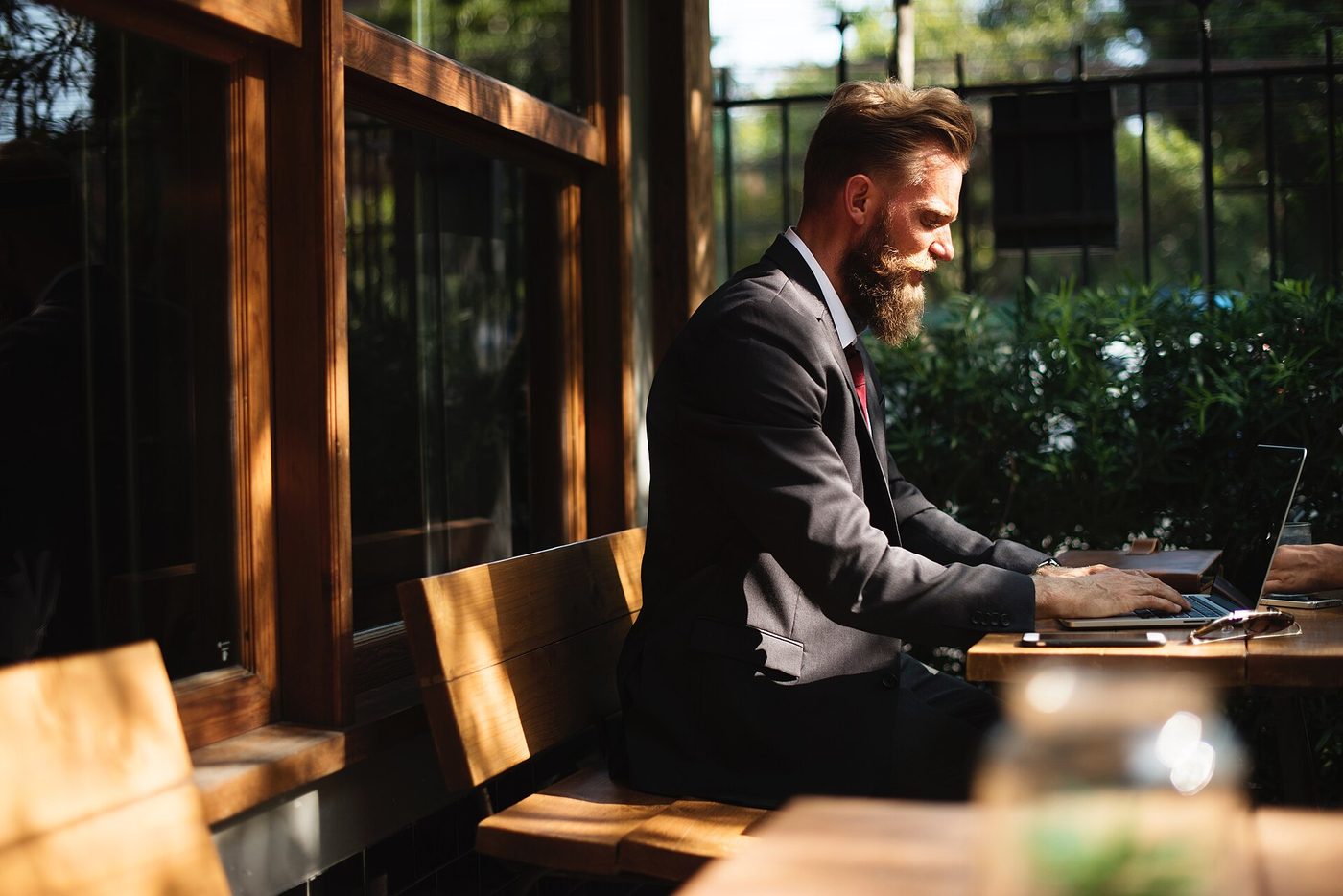 Man working on a laptop at a sunlit open-air cafe table surrounded by greenery