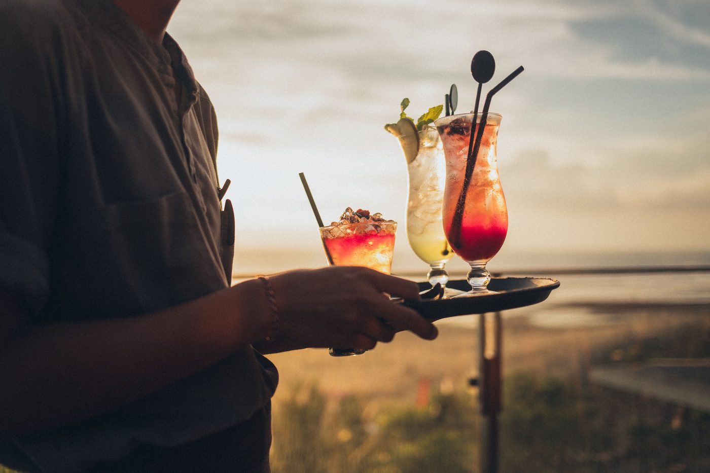 Silhouetted server carrying a tray of tropical cocktails at a sunset beach bar