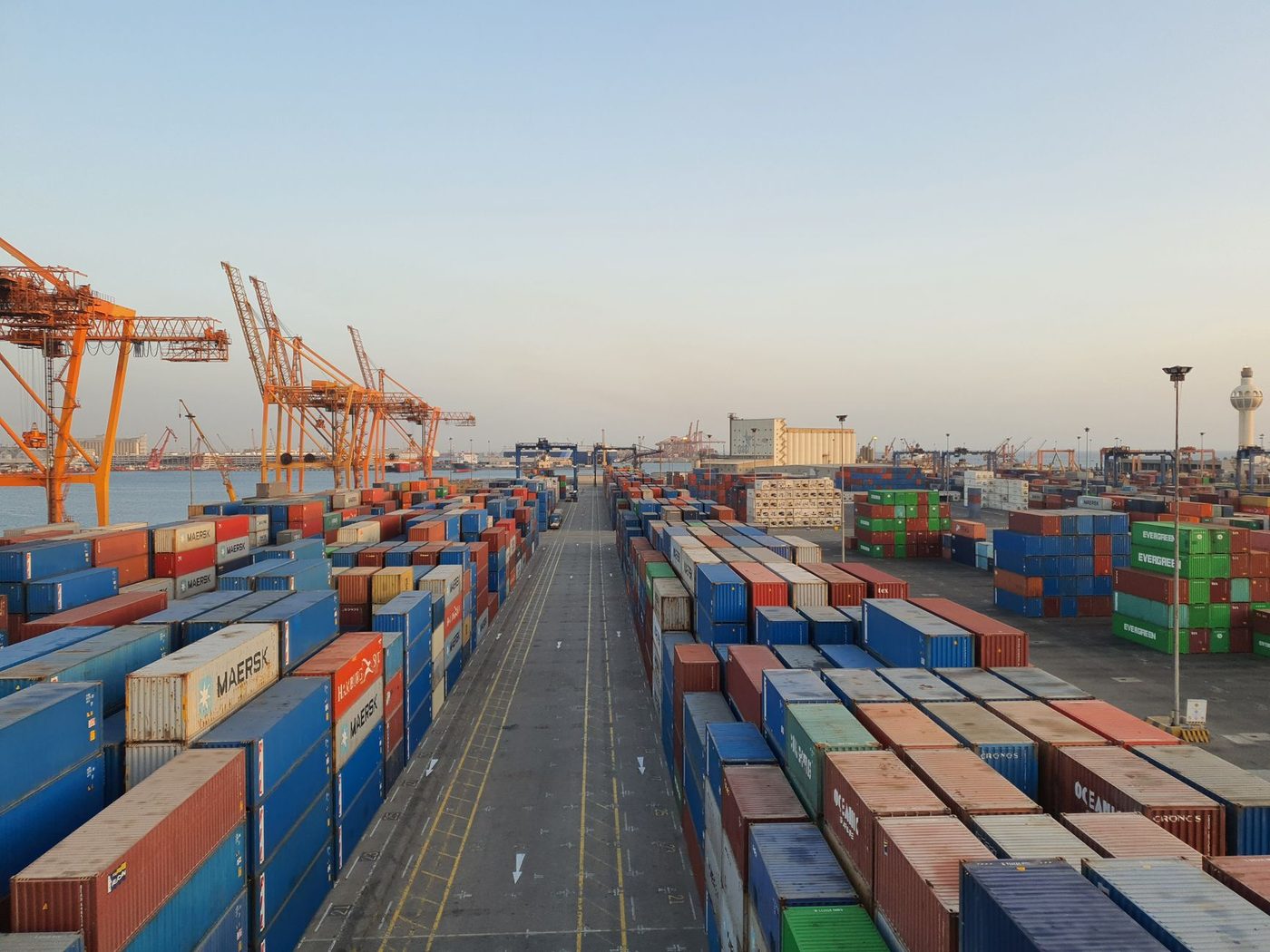 Rows of stacked shipping containers and cranes at a busy cargo port at sunrise