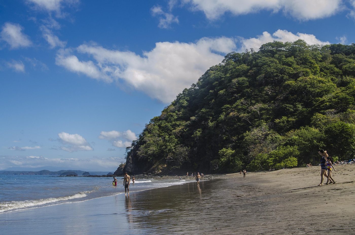 Beachgoers walk the tide line below a forested headland at Playa Matapalo, Guanacaste