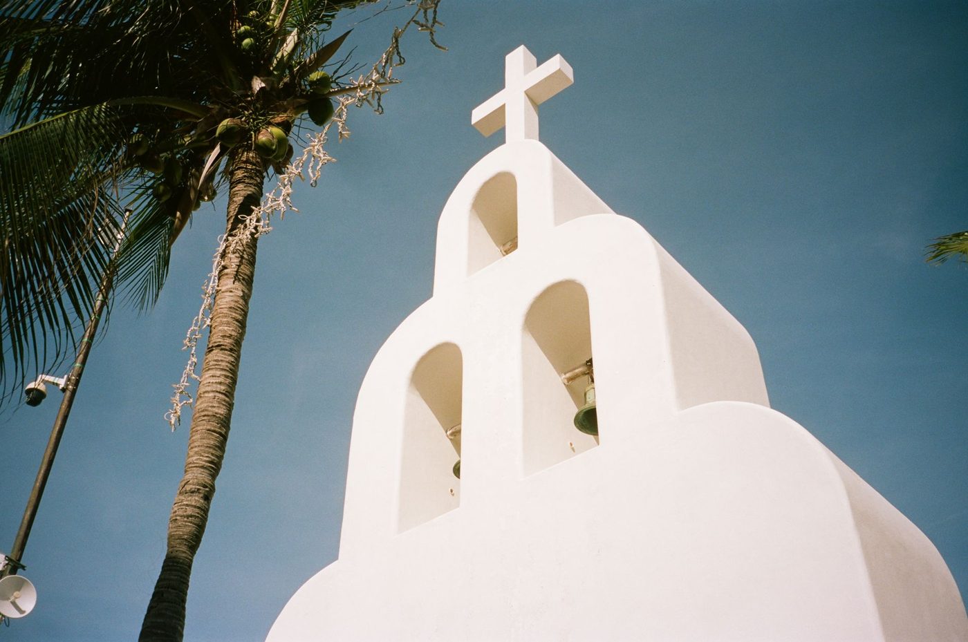 Simple white church bell tower with cross against blue sky and palm tree