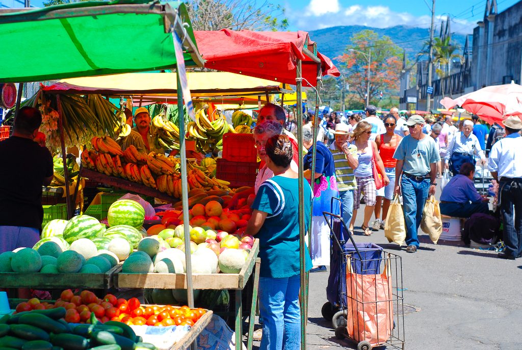 Shoppers browsing watermelons, bananas, and tomatoes at a Costa Rican farmers market