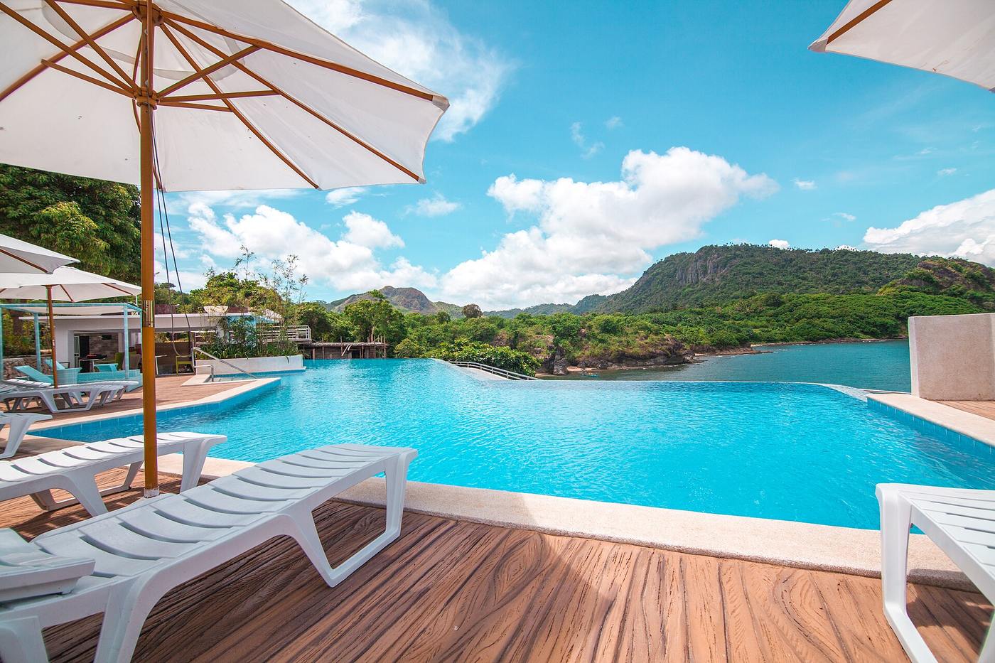 Infinity pool with lounge chairs overlooking forested hills and a quiet tropical bay